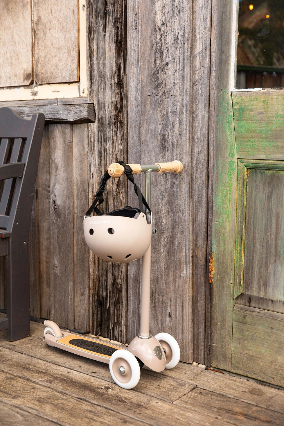 Banwood vintažinis 3-ratis paspirtukas su krepšeliu ir šalmas, stovintis prie medinės sienos.