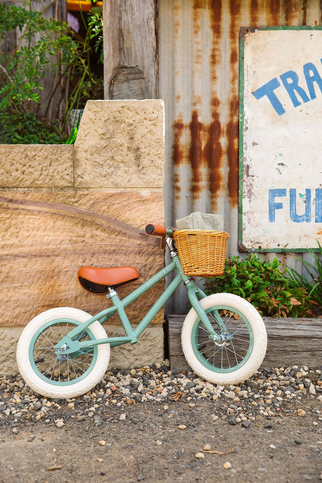 Banwood vintažinis balansinis dviratis First Go - Forest su pintu krepšiu ir šviesiais ratais, stovintis lauke.