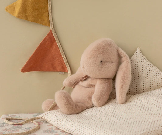 Maileg pliušinis žaislas - triušiukas sėdi ant pagalvėlės šalia spalvotų vėliavėlių.